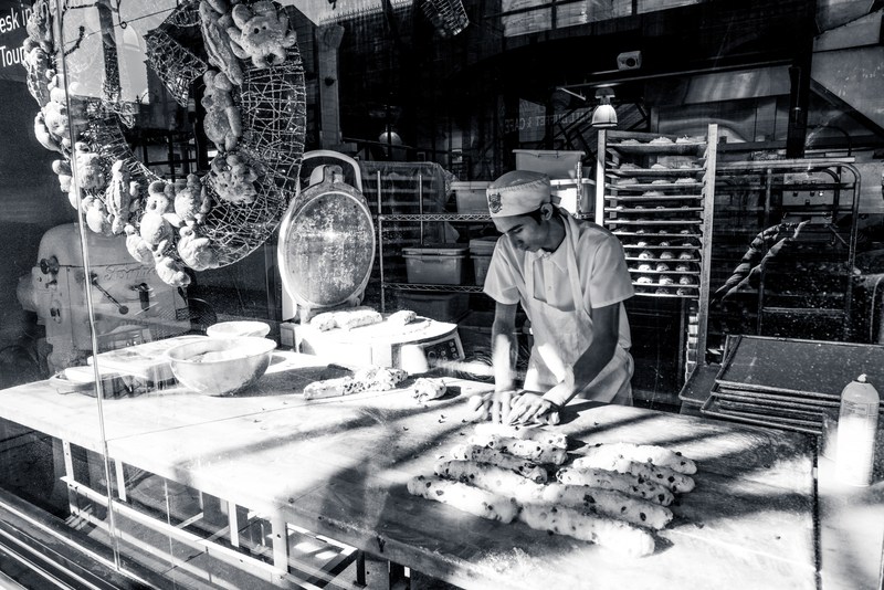 A baker works with dough behind reflective glass in a monochrome shop interior.