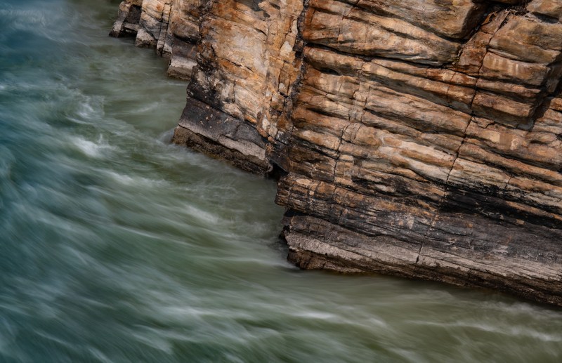 Horizontal ochre rock layers meet green water flowing around the base of a cliff.