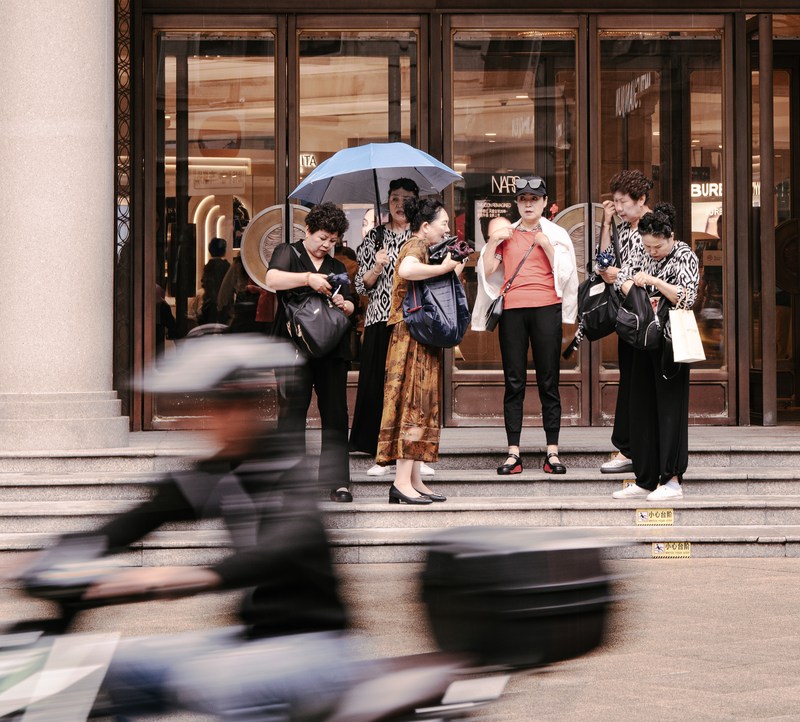 A group pauses on shop steps while a scooter crosses the foreground in a blur.