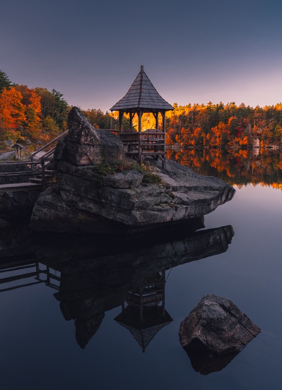 An empty pavilion frames sunlit autumn trees beside a still lake at Mohonk in the morning.