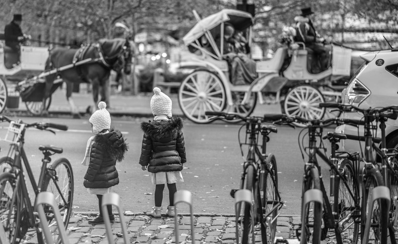 Two children in matching knit hats and dark coats watch horse carriages from between parked bicycles, in black and white.